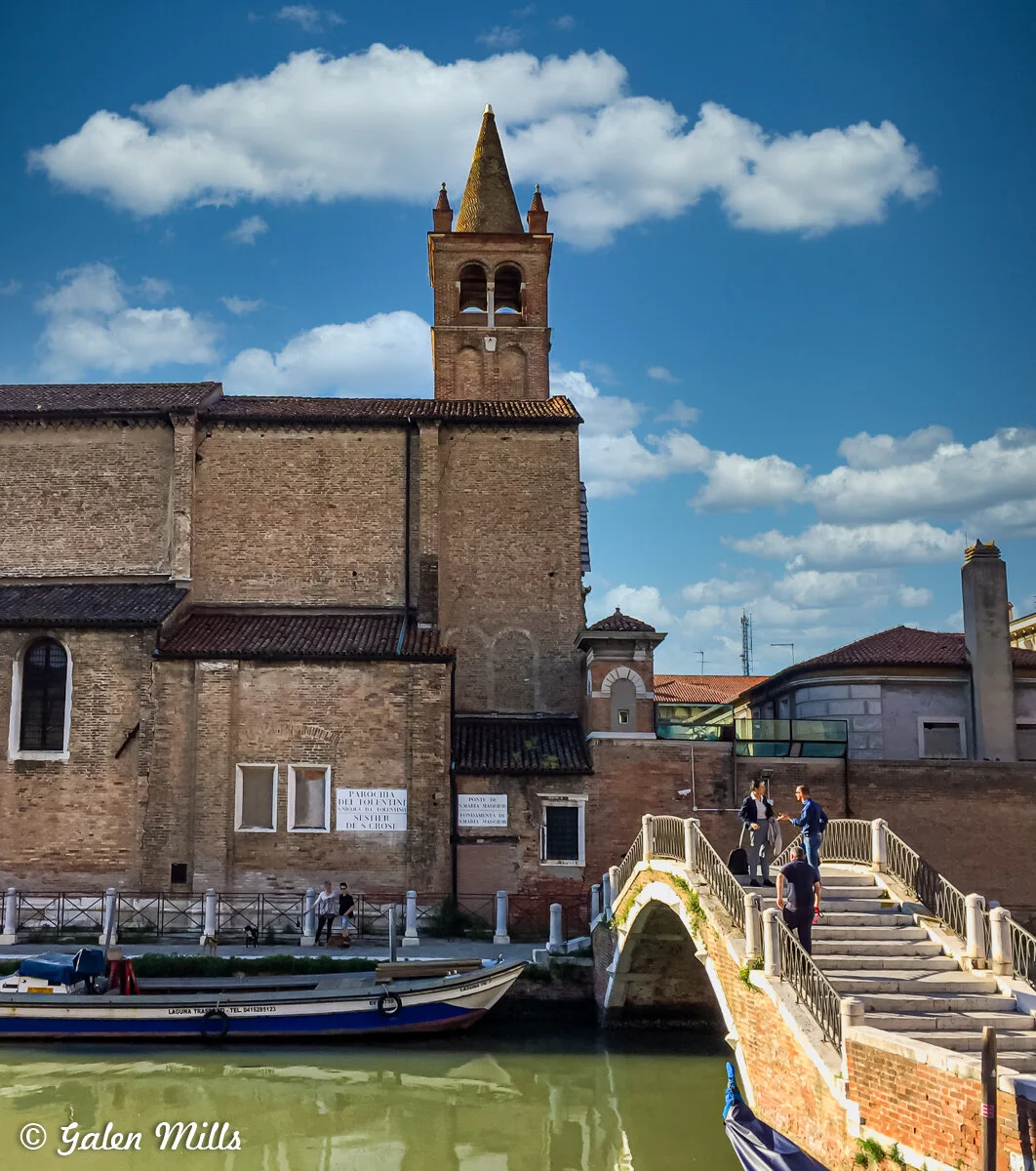 Historic building and bridge in Venice with people crossing, canal with boats, and blue sky with clouds.