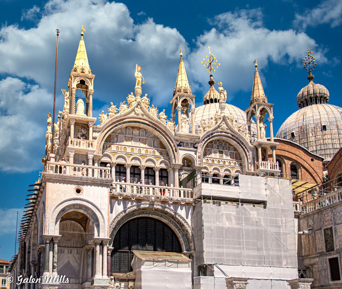 Facade of St. Mark's Basilica with ornate arches, golden spires, and sculptures under a blue sky with clouds, partial restoration work visible.