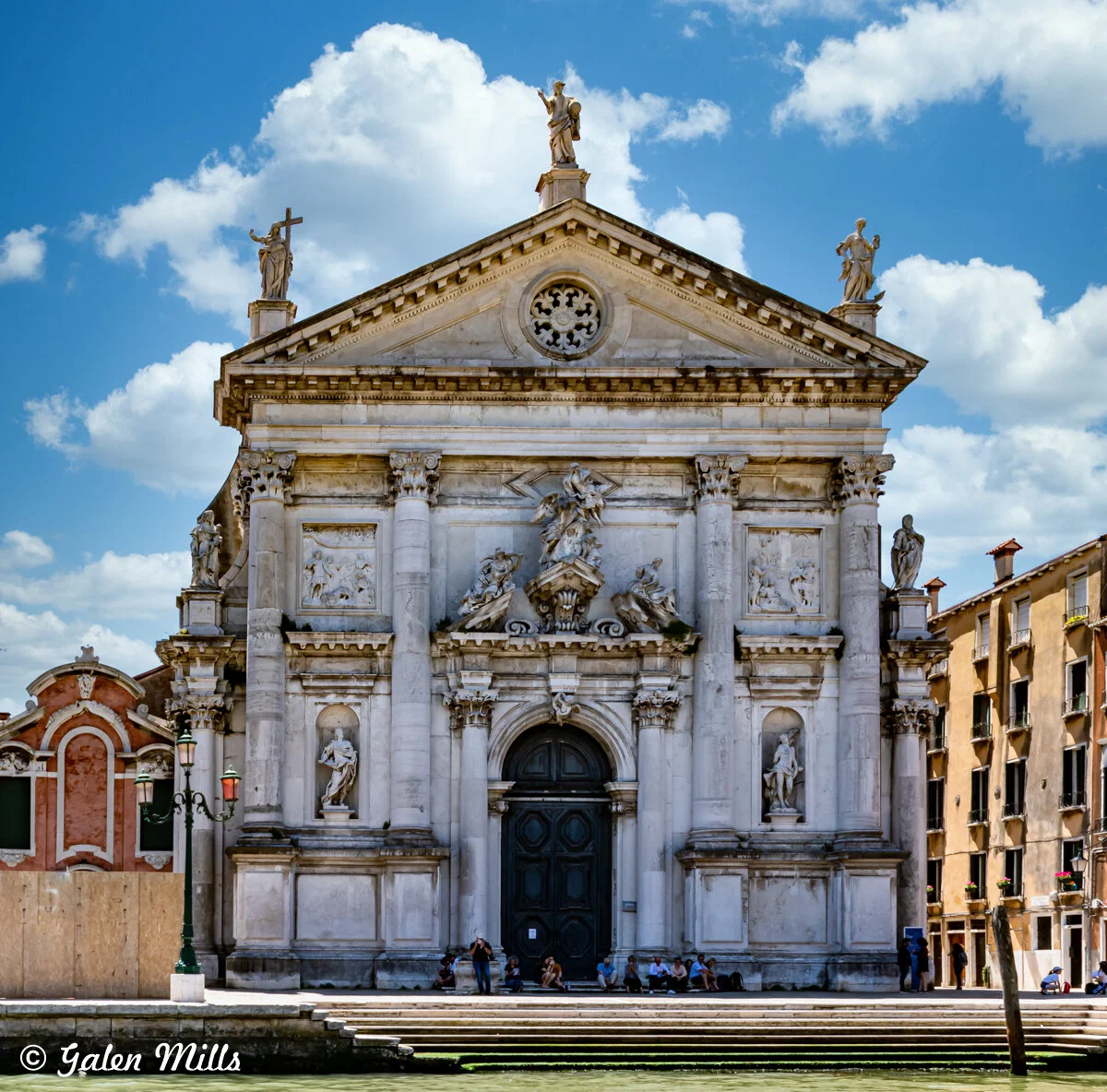 Facade of the Church of San Stae in Venice, Italy, featuring ornate sculptures and architectural details, under a blue sky with clouds.