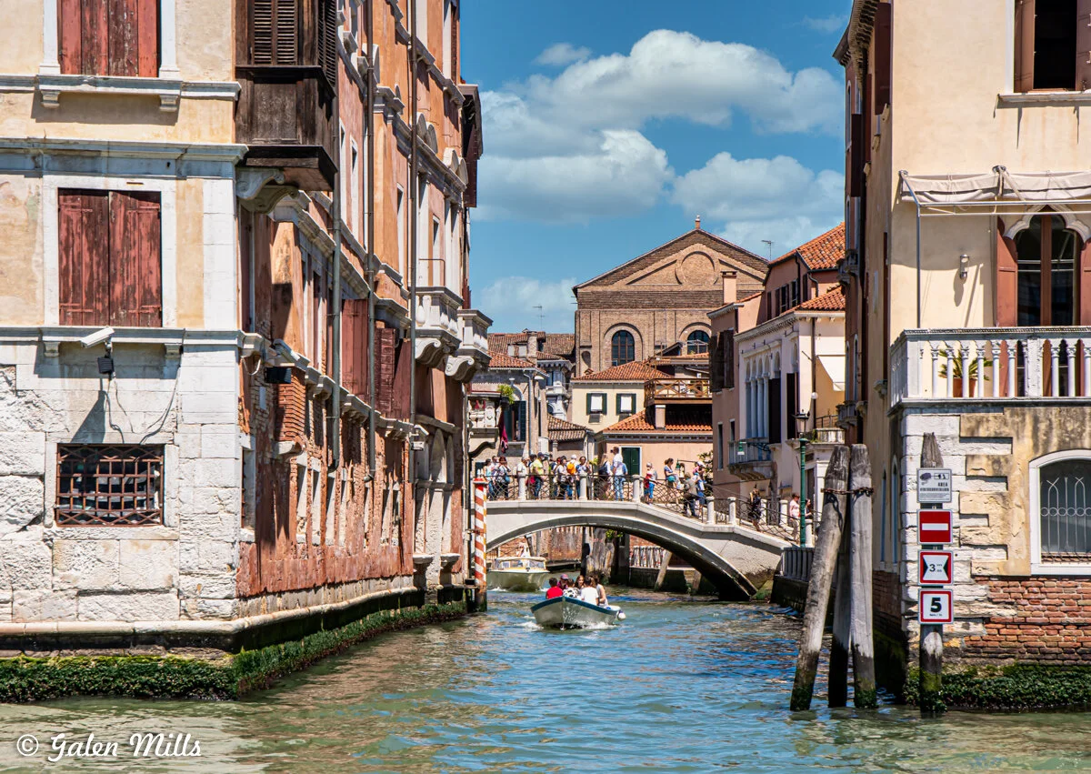 Venetian canal with a boat, historic buildings, and a stone bridge crowded with tourists under a blue sky.