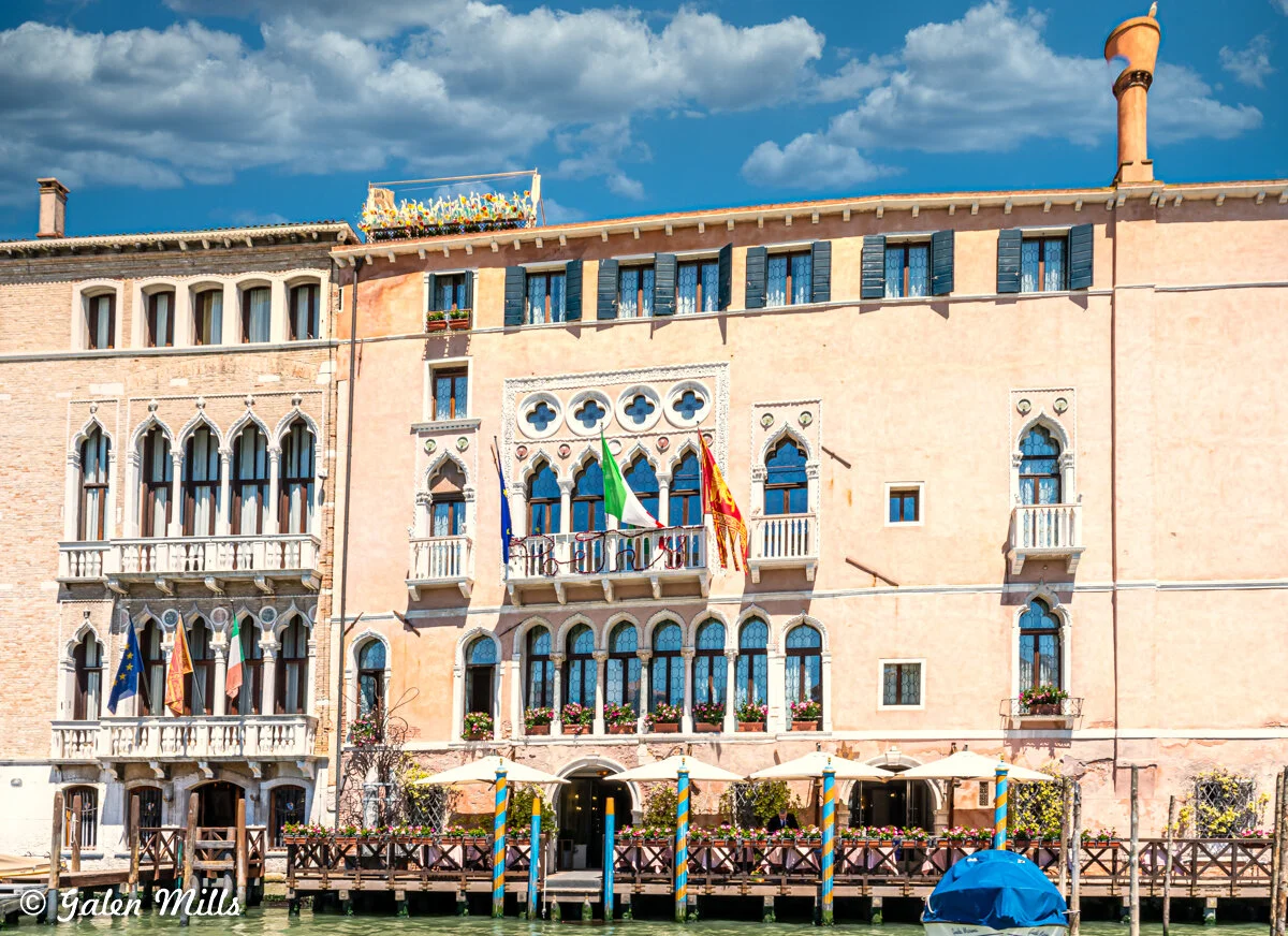 Historic Venetian building with arched windows and balconies, adorned with flags, beside a canal under a sunny sky.