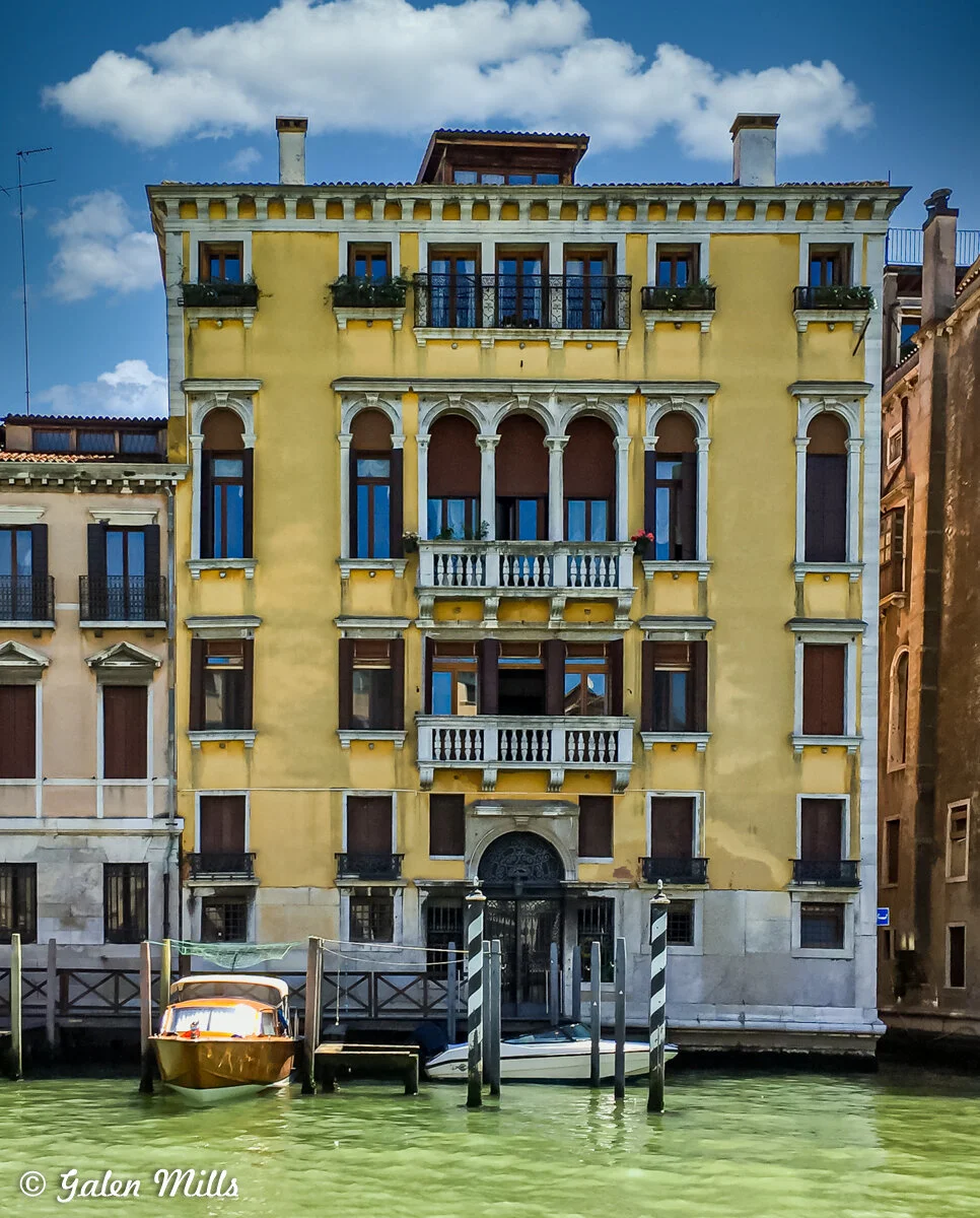 Yellow Venetian building with arched windows overlooking a canal, featuring a dock with a wooden boat moored in front.