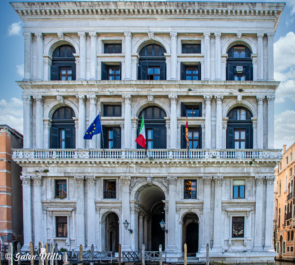 Historic European-style building with arched windows, European Union, Italian, and another flag displayed on a balcony, featuring classical columns and decorative details.
