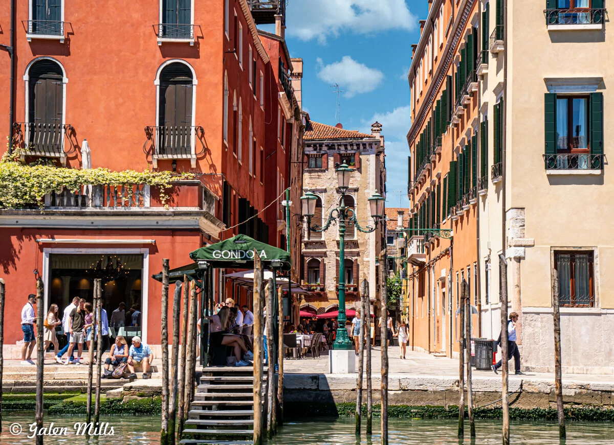 Colorful buildings and a gondola dock along a canal in Venice, Italy, with people walking and sitting nearby.