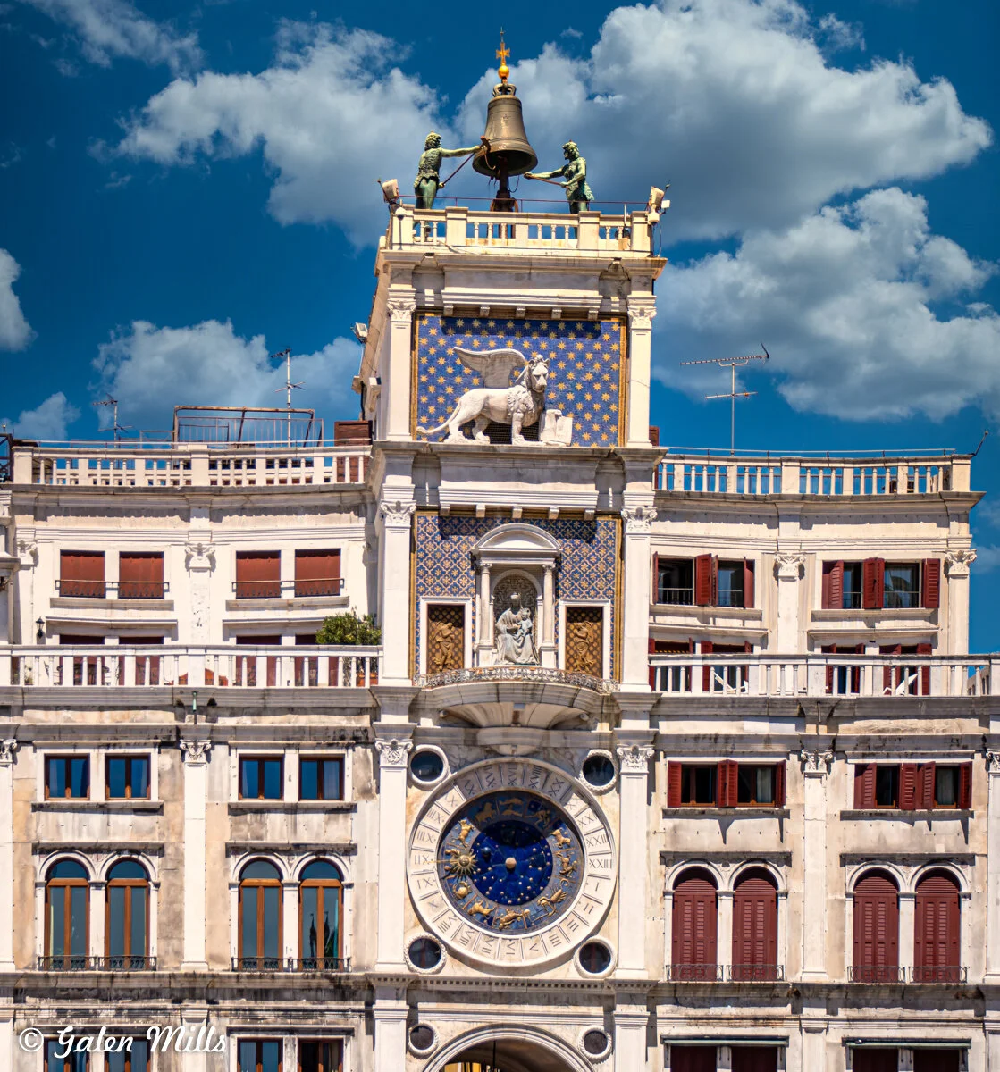 Close-up of St. Mark's Clocktower in Venice, featuring a large astronomical clock and a bell with statues on top, against a blue sky with clouds.
