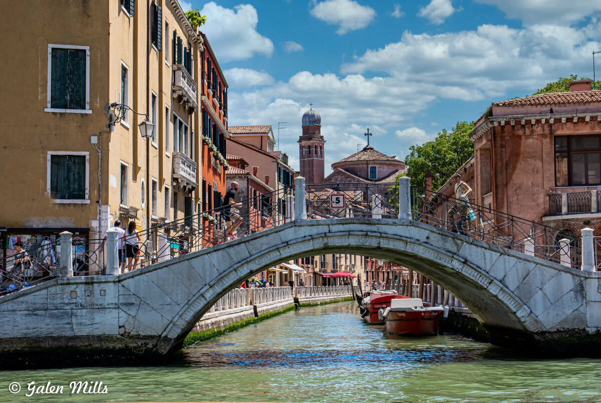 View of a canal in Venice, Italy, with a stone bridge crossing over and colorful buildings lining the waterway. Several people are walking on the bridge, and boats are moored along the canal. The sky is blue with scattered clouds.