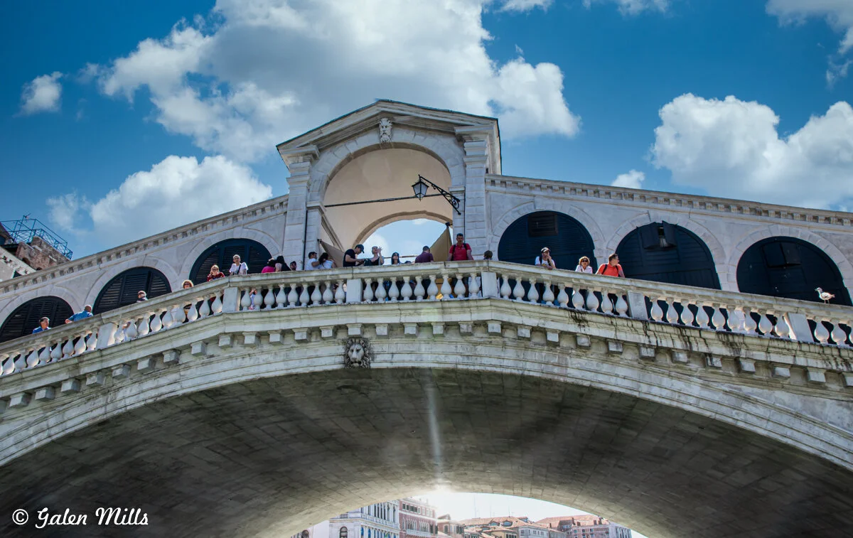 View of the Rialto Bridge in Venice, Italy, with tourists standing on the bridge under a blue sky with clouds.