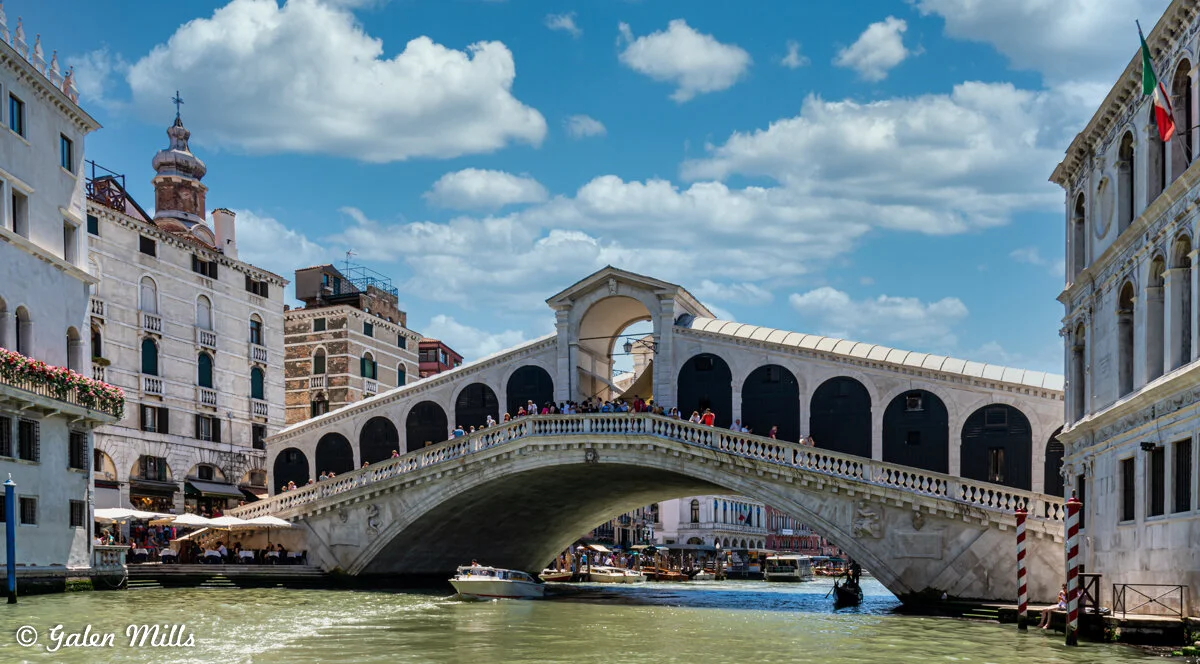 Rialto Bridge spanning the Grand Canal in Venice under a blue sky with white clouds, surrounded by historic buildings and gondolas on the water.