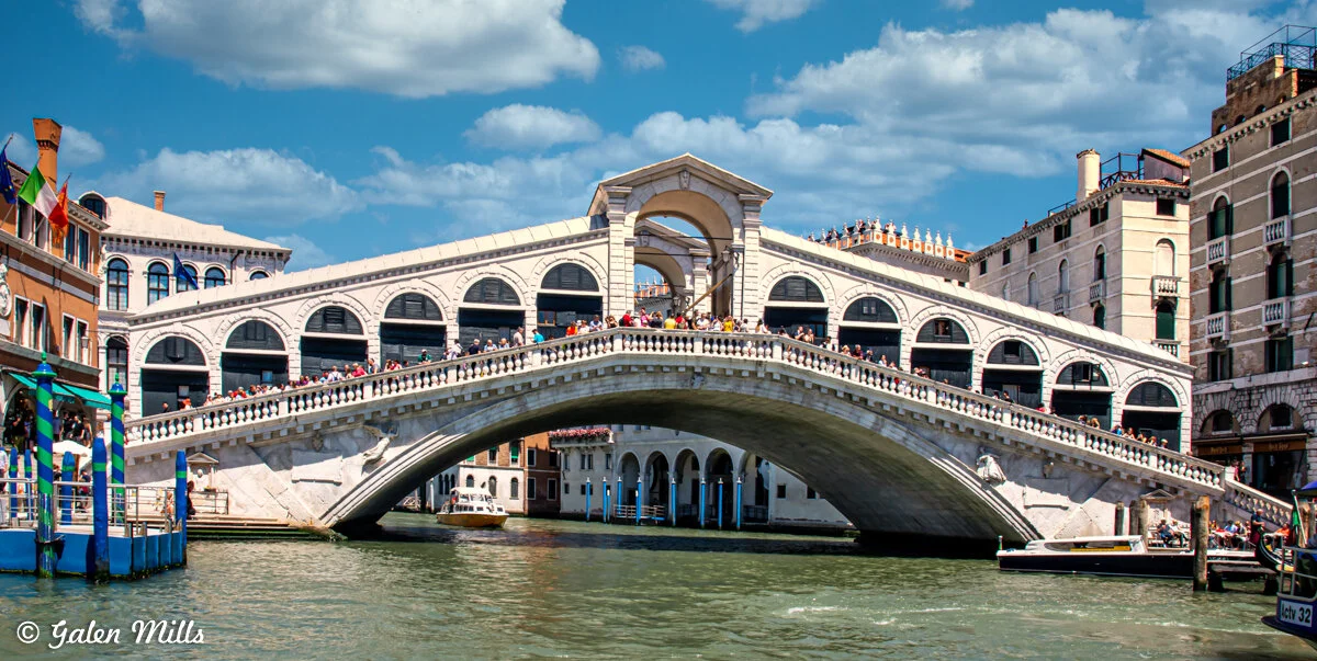 Rialto Bridge in Venice, Italy, with tourists walking on it and canal boats below, set against a blue sky with clouds.