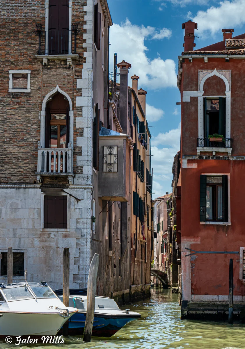 A narrow canal in Venice, Italy, flanked by two old buildings with arched windows and shutters. The canal has two small boats docked and is set against a blue sky with clouds.