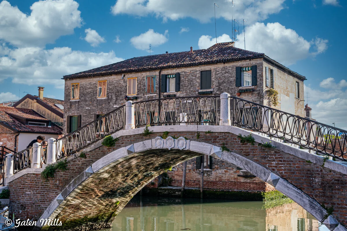 Historic brick bridge with iron railing over canal in front of old stone buildings with shuttered windows, under a blue sky with clouds.