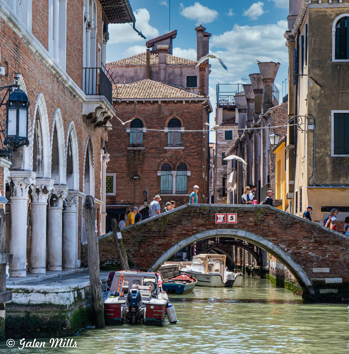 Venice canal with a brick bridge, lined with boats and historic buildings in the background. People walking on the bridge under a blue sky with scattered clouds.