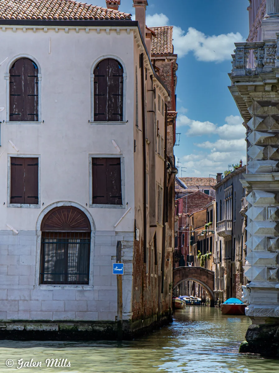 Narrow canal in Venice, Italy, flanked by historic buildings with shuttered windows. A small bridge arches over the canal in the background, and the sky is clear with few clouds.