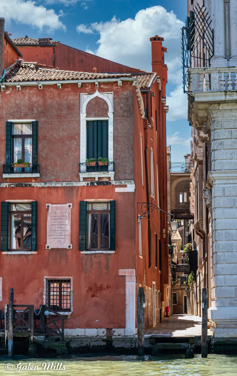Scenic view of a red Venetian building alongside a canal with a small pier and arched windows, next to a narrow alleyway between buildings. Bright blue sky with clouds overhead.