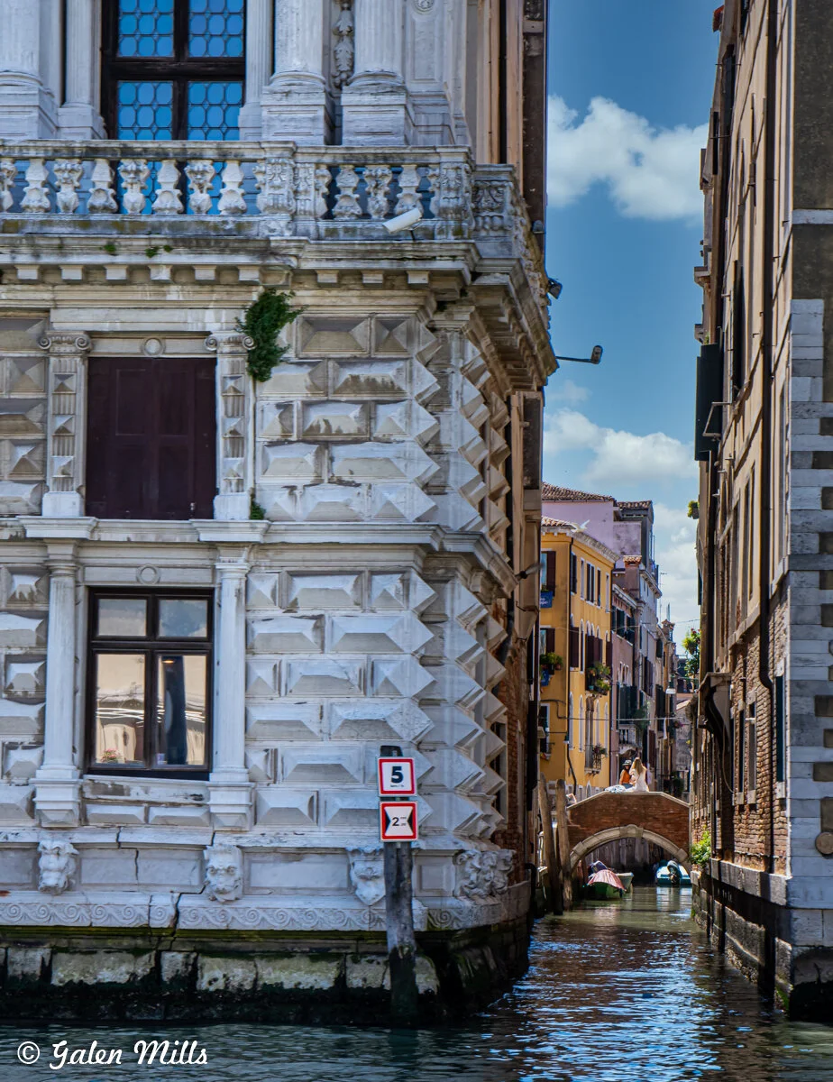 View of a Venetian canal with a historic stone building featuring decorative architecture on the left. A narrow waterway runs between buildings, leading to a small arched bridge. Two signs with speed limits are visible. The scene includes calm water 