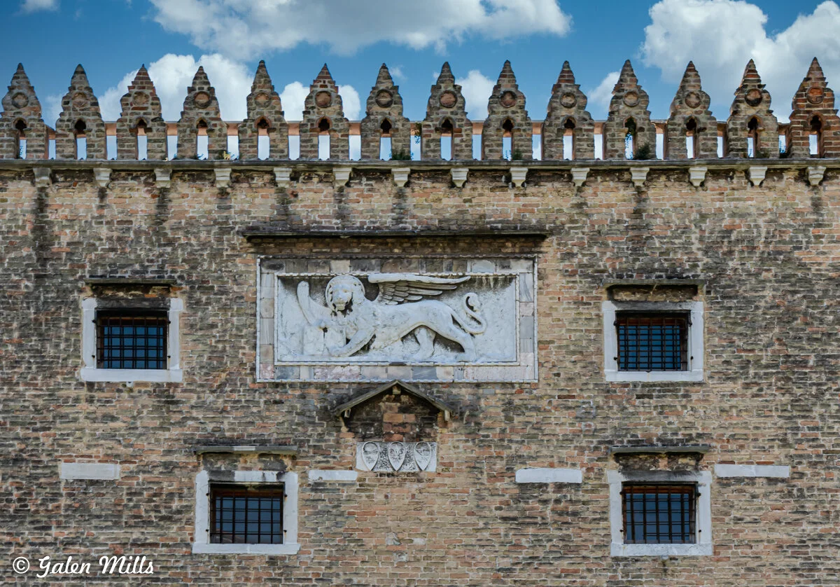 Brick building facade with a marble relief of a winged lion and decorative crenellations, featuring barred windows and a cloudy sky background.