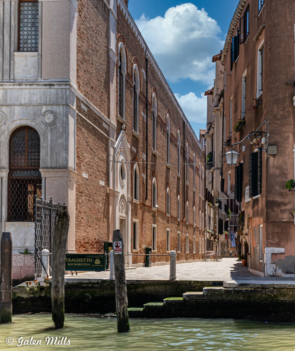 Venetian canal scene with historic buildings; brick structure on left, narrow alley with windows and laundry, green water in foreground, blue sky with clouds.