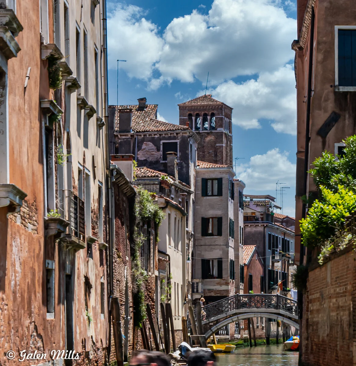 Narrow canal in Venice with historic buildings, a small bridge, and a boat. Cloudy blue sky.