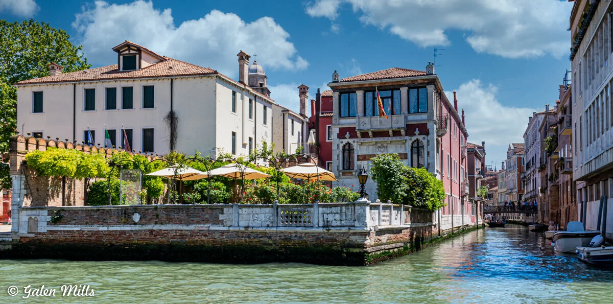 Venice canal scene with historic buildings, outdoor cafe, and blue sky.