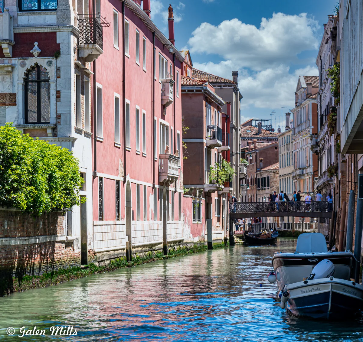 Venetian canal with colorful buildings, boats on water, and a pedestrian bridge with people.