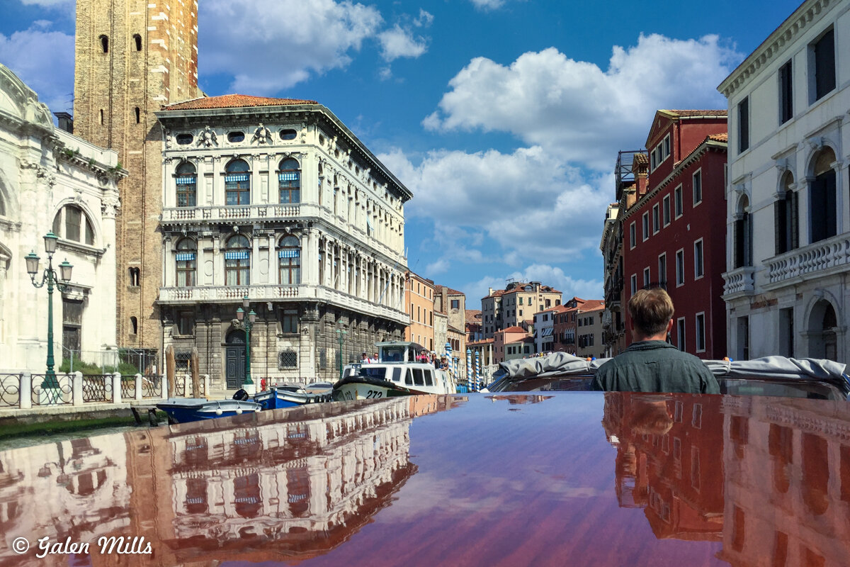 Scenic view of a canal in Venice, Italy, featuring historic buildings, a boat in the foreground, and a person looking towards the buildings under a blue sky with clouds.