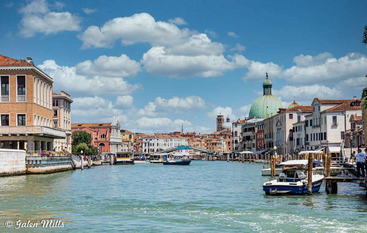 Venice Grand Canal with boats, colorful buildings, and a church dome under a blue sky with clouds.