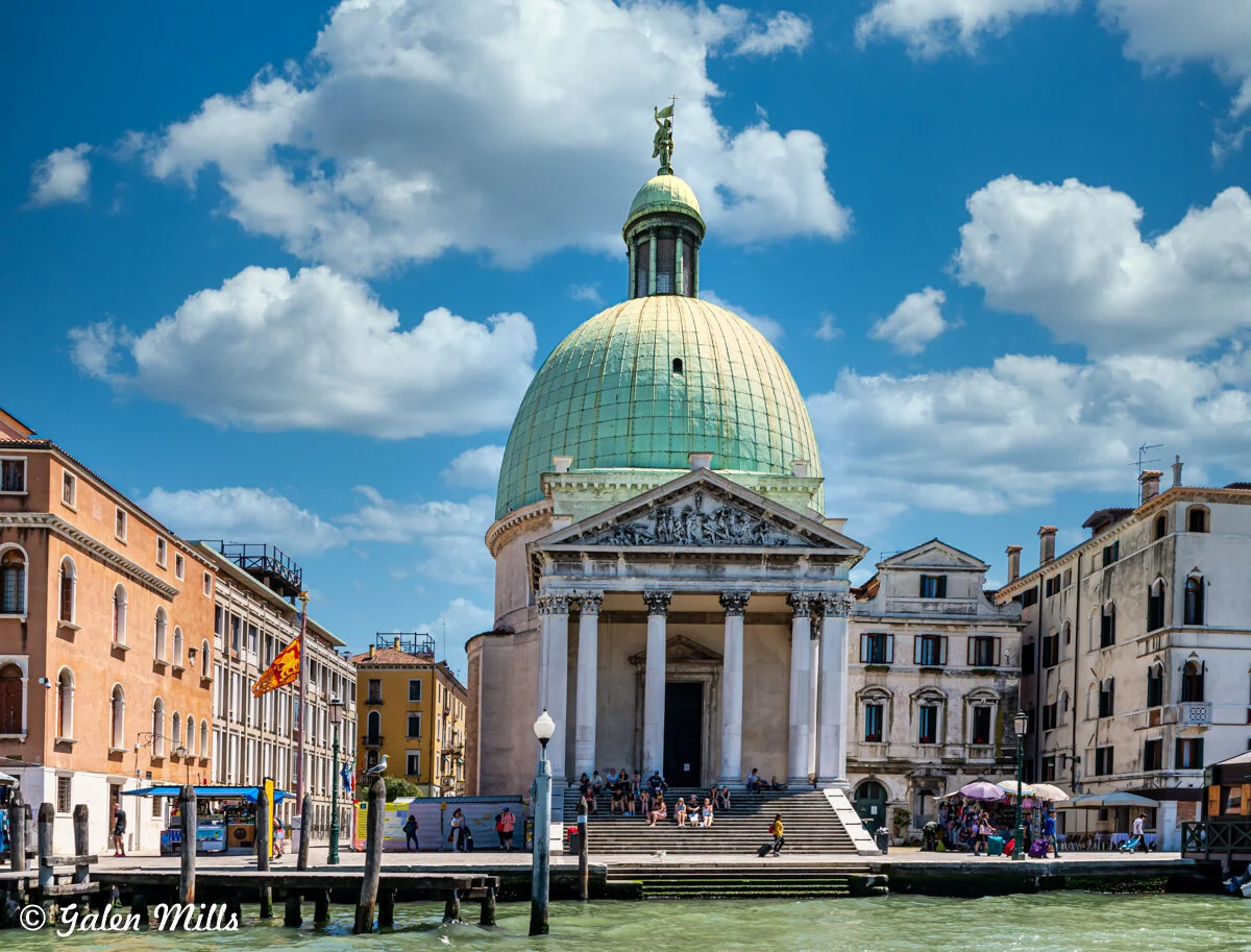Grand building with large green dome and classical columns, set in a European cityscape by a canal, under a blue sky with clouds.