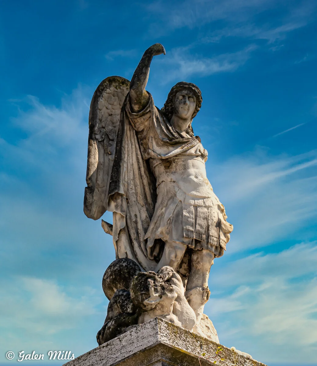 Marble statue of an angel with wings and raised arm against blue sky.