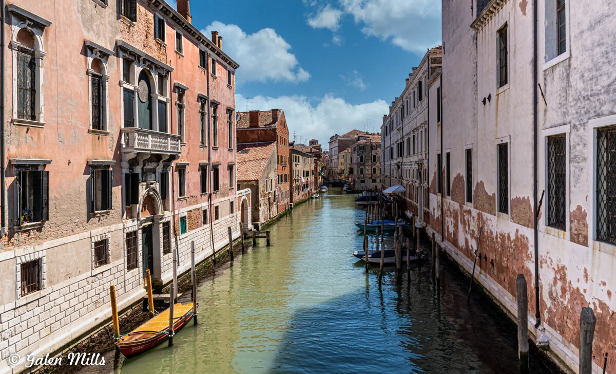 A narrow canal in Venice surrounded by old buildings with balconies and shutters, reflecting in the water; gondolas and mooring posts are visible, under a blue sky with clouds.