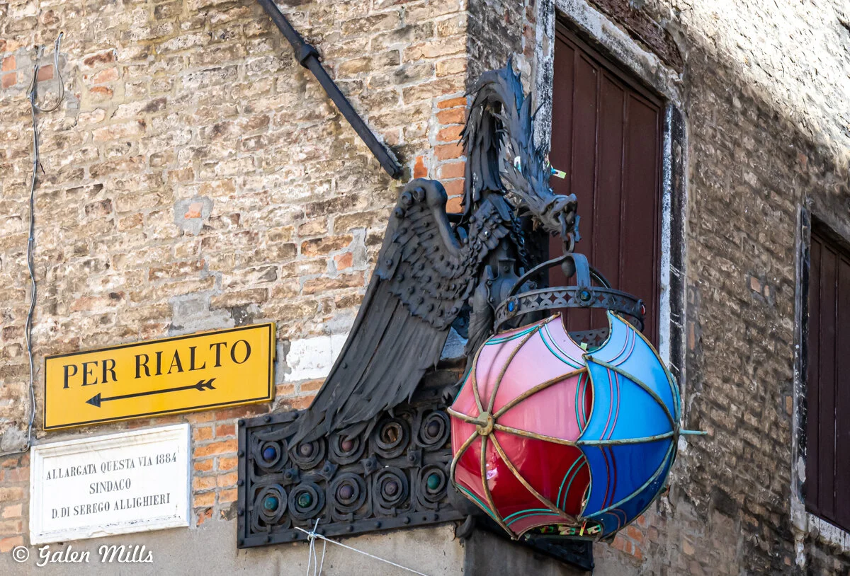 Dragon relief and colorful lamp on historic brick wall next to "Per Rialto" directional sign in Venice.