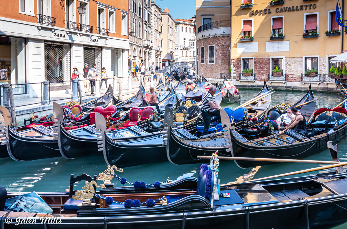 Gondolas in a canal in Venice, Italy, with people walking along the waterfront and a hotel in the background.