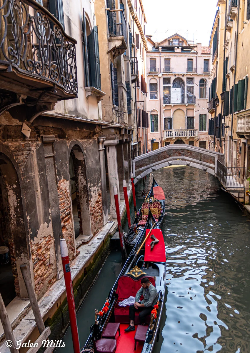 Narrow canal in Venice with gondolas, stone bridge, and old buildings.