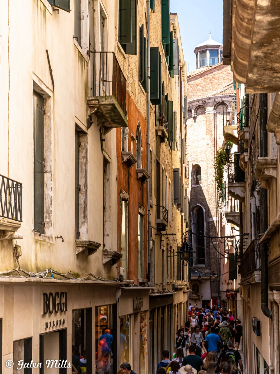 Narrow alley in Venice with people walking, surrounded by historic buildings and a visible church tower.