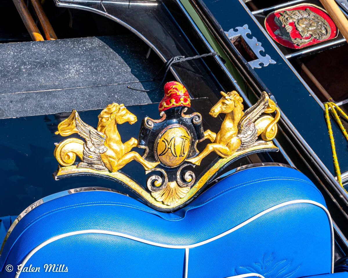 Close-up of a decorative emblem on a gondola, featuring gold pegasi and a crest with a red and black design, against a blue seat.