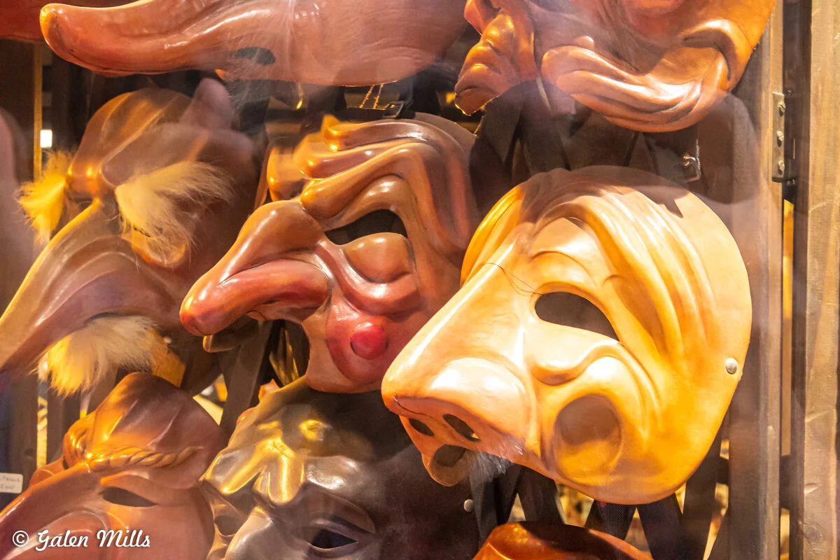 A display of various theatrical masks with exaggerated features, including long noses and pronounced facial expressions. The masks are made of wood or a wood-like material.