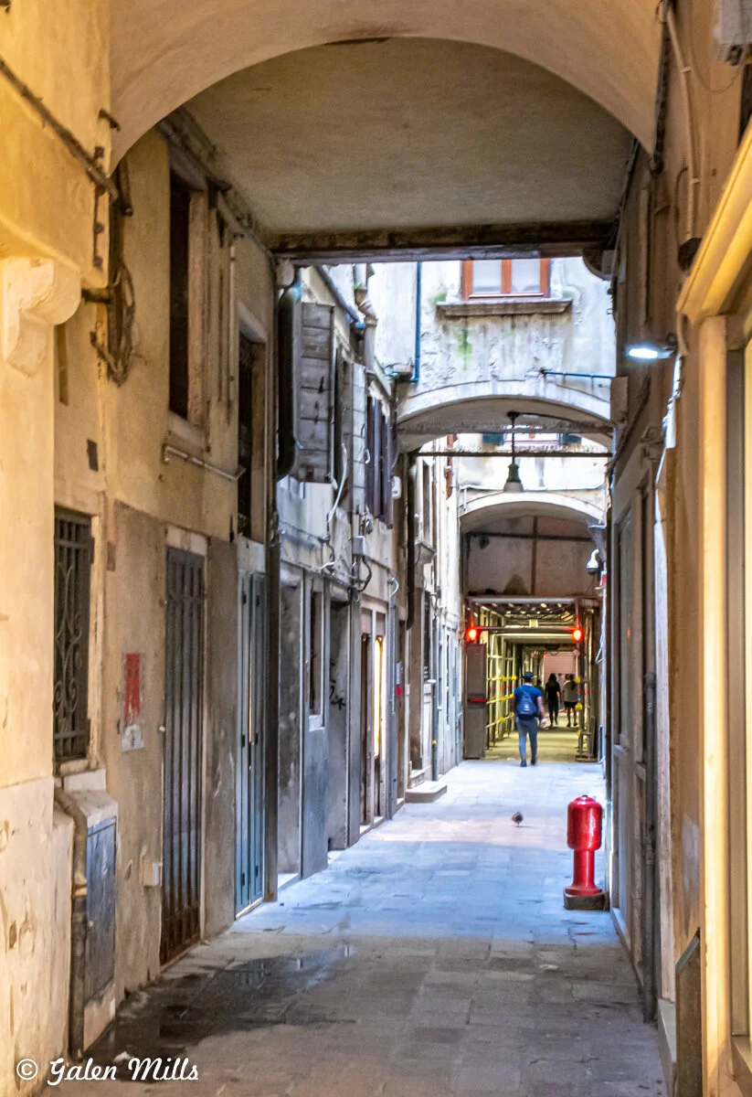 Narrow alleyway with archways, old buildings, and a red bollard. A person walking in the distance.