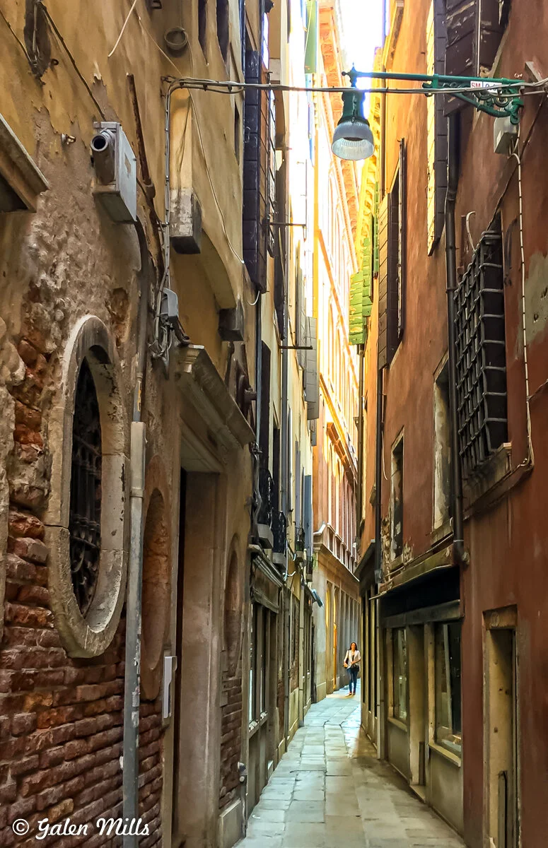 Narrow alleyway in Venice with person walking, surrounded by old buildings.