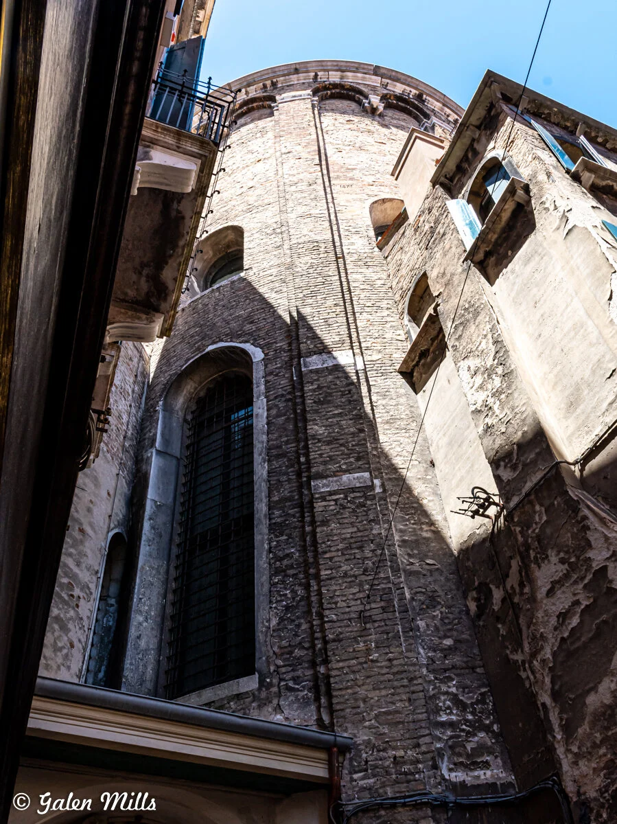 Narrow alleyway with tall brick buildings, featuring arched windows and balconies, under a clear blue sky.