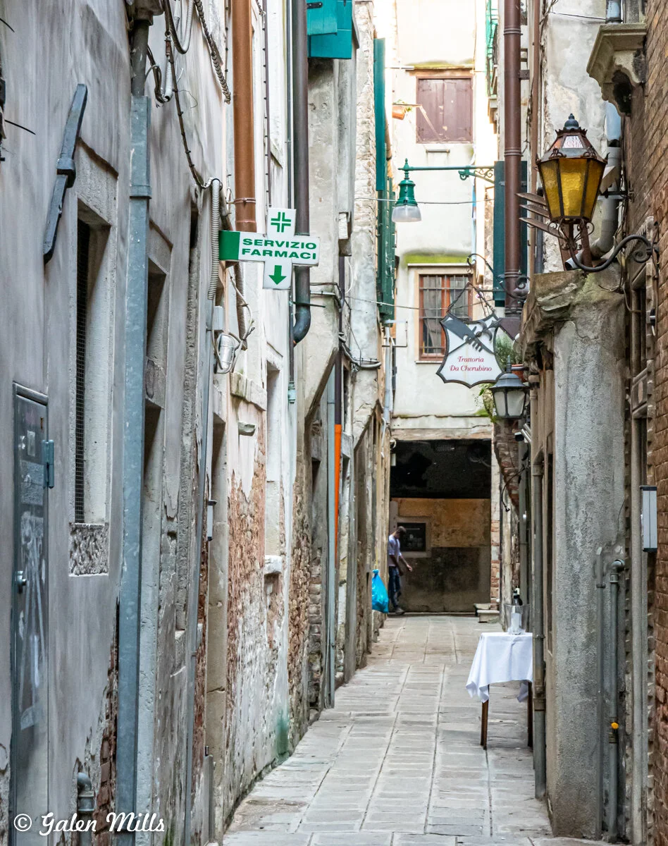 Narrow alleyway in Venice with weathered buildings, cobblestone path, pharmacy sign, and restaurant table.