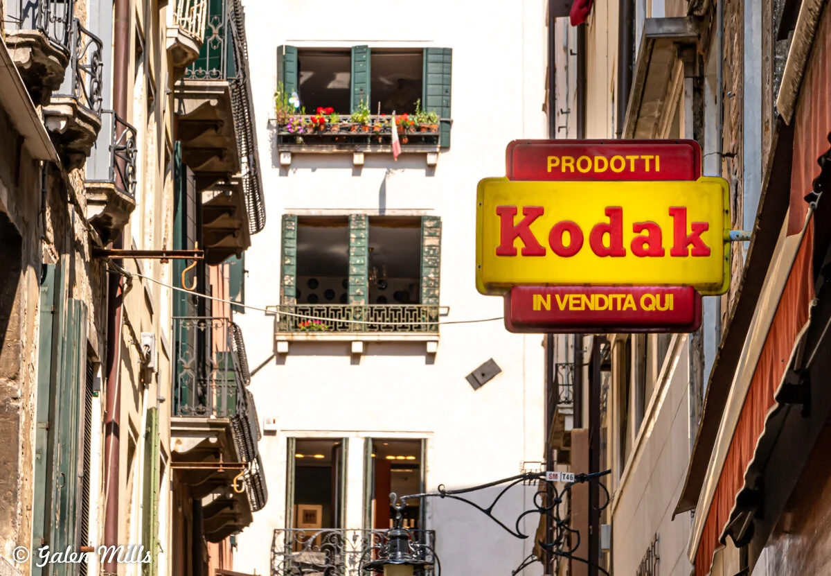 Narrow European street with a yellow and red Kodak sign, buildings with balconies and flowers, sunny day.