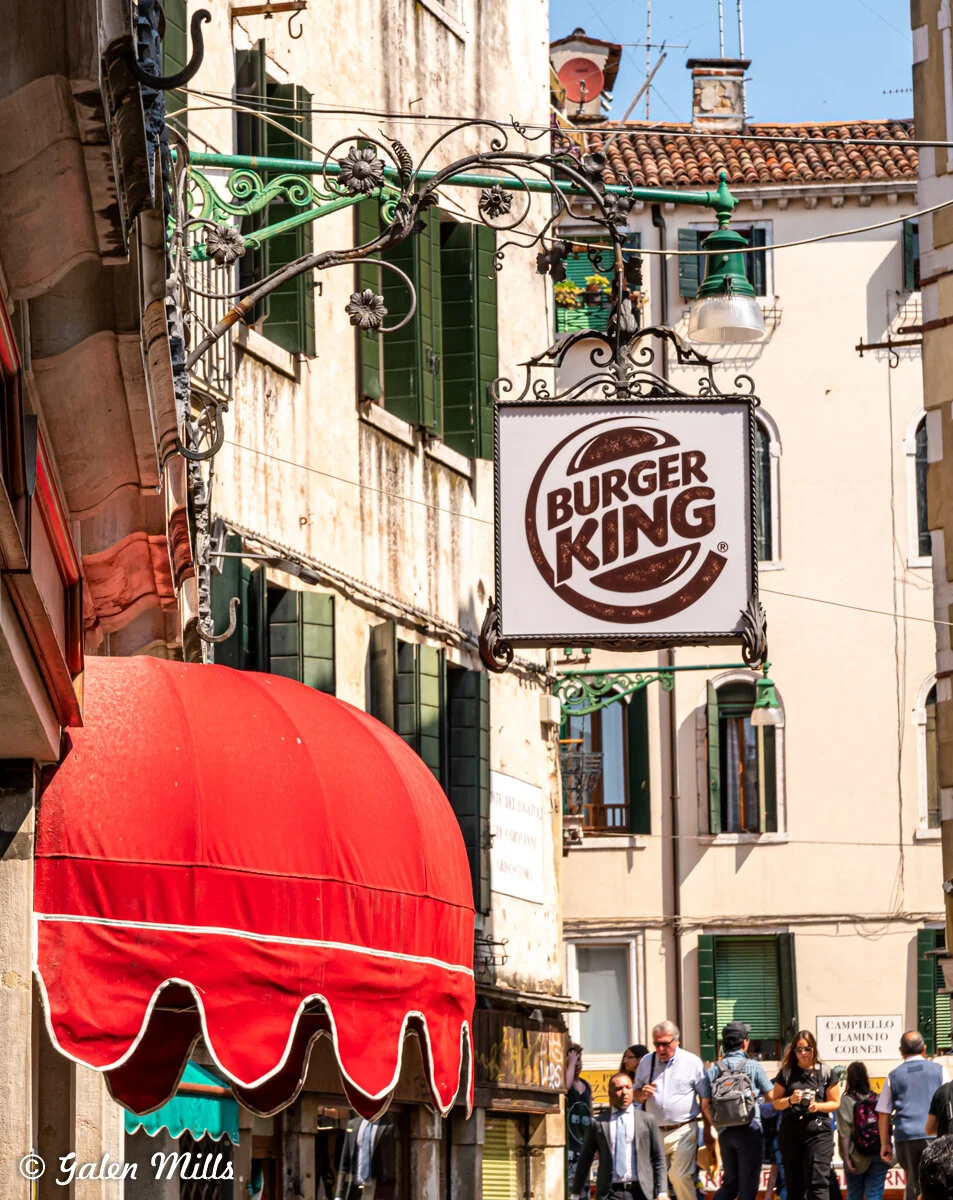 Street view featuring a Burger King sign, a red awning, and buildings with green shutters, with people walking below.