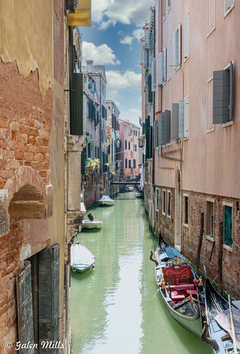 Narrow Venetian canal with boats and gondolas, surrounded by pastel-colored buildings, under a partly cloudy sky.