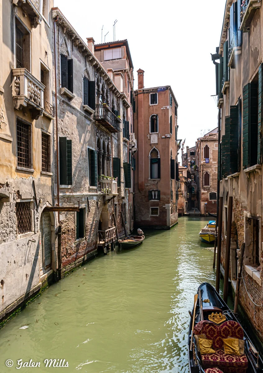 A narrow canal in Venice, Italy, flanked by old buildings with balconies and shutters. A gondola with ornate seating and a small motorboat are moored along the canal's edge.