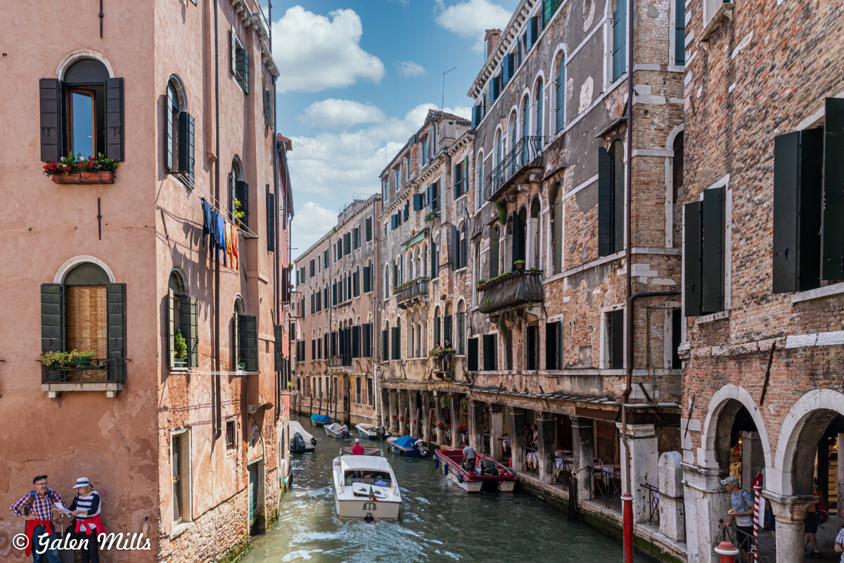 Scenic view of a canal in Venice, Italy, with colorful buildings, open windows, and boats on the water. People visible near the canal, blue sky with clouds above.