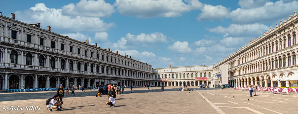 St. Mark's Square in Venice with people walking, pigeons, and historic buildings under a blue sky.