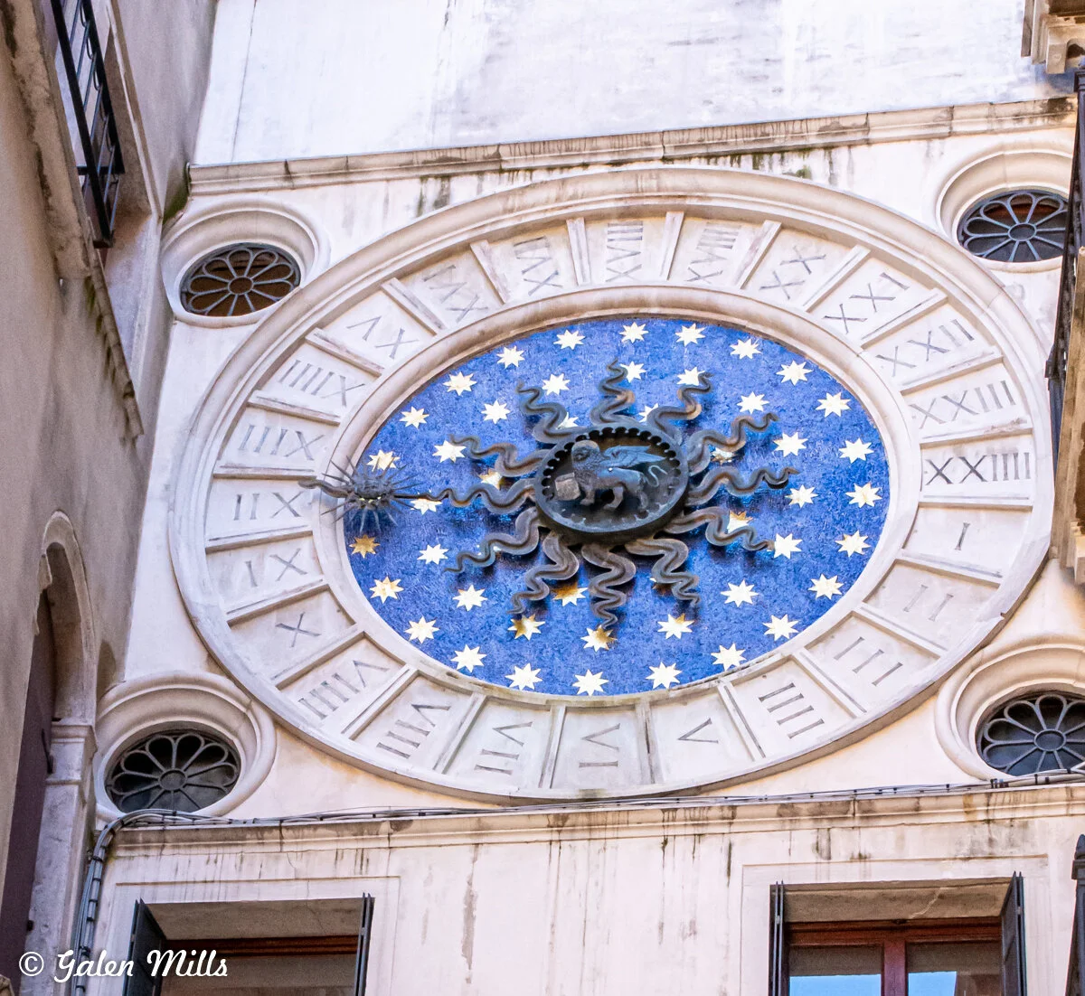 Circular astronomical clock with Roman numerals and a central blue starry background, featuring a sunburst and a lion emblem.
