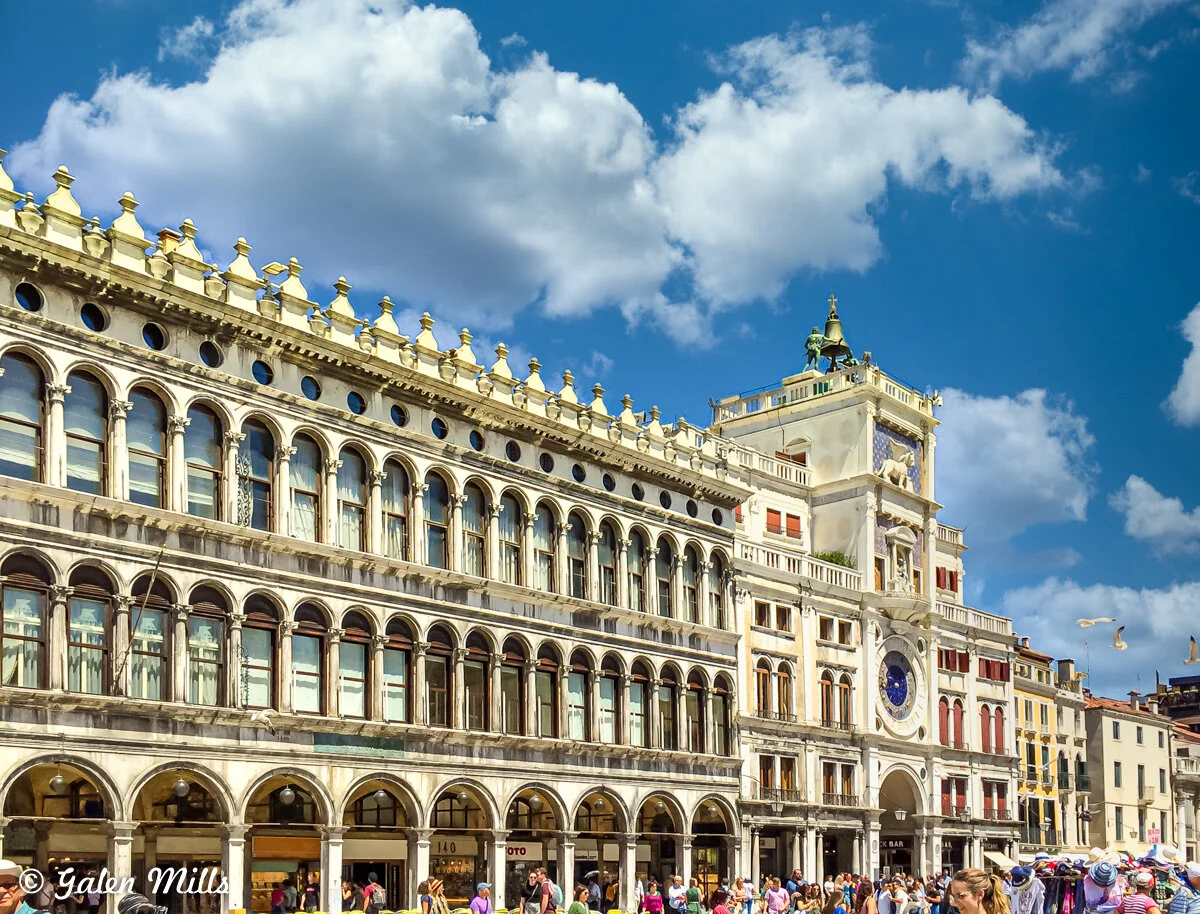 Historic building with arched windows and ornate facade in St. Mark's Square, Venice, under a blue sky with clouds.