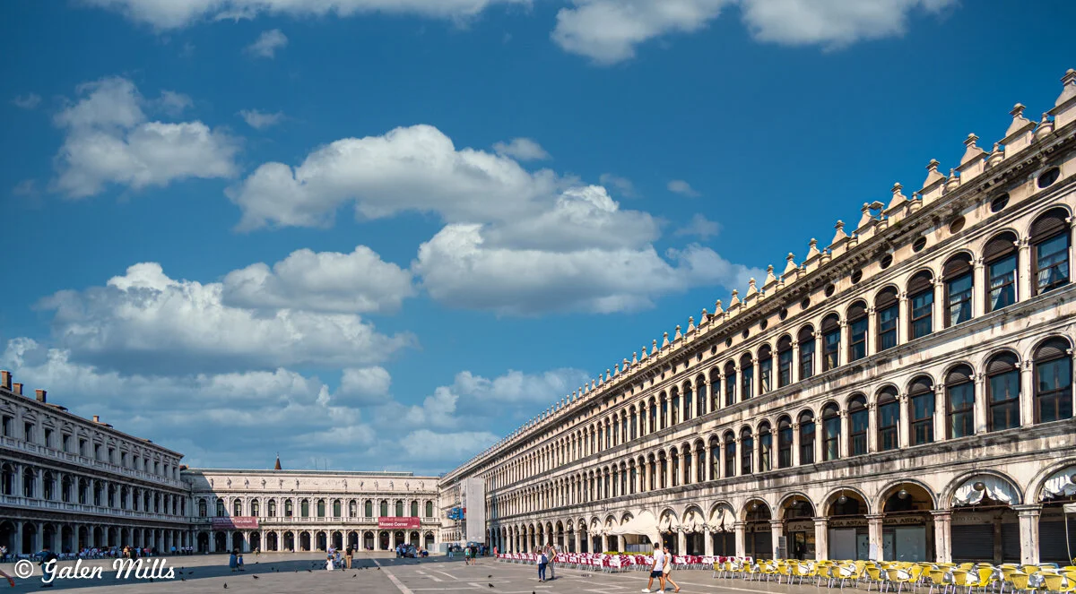 St. Mark's Square, Venice, under a clear blue sky with white clouds, featuring elegant arched windows and yellow chairs in front of the buildings.