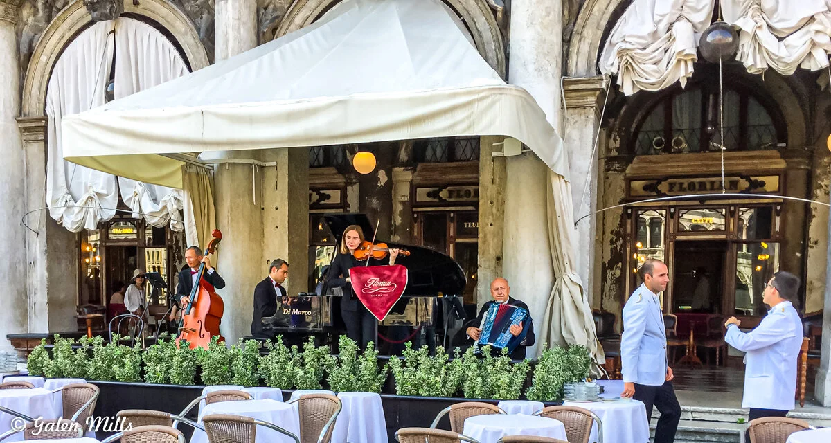 Outdoor cafe with live musicians playing violin, accordion, and piano; waitstaff in white jackets; tables set with white tablecloths.