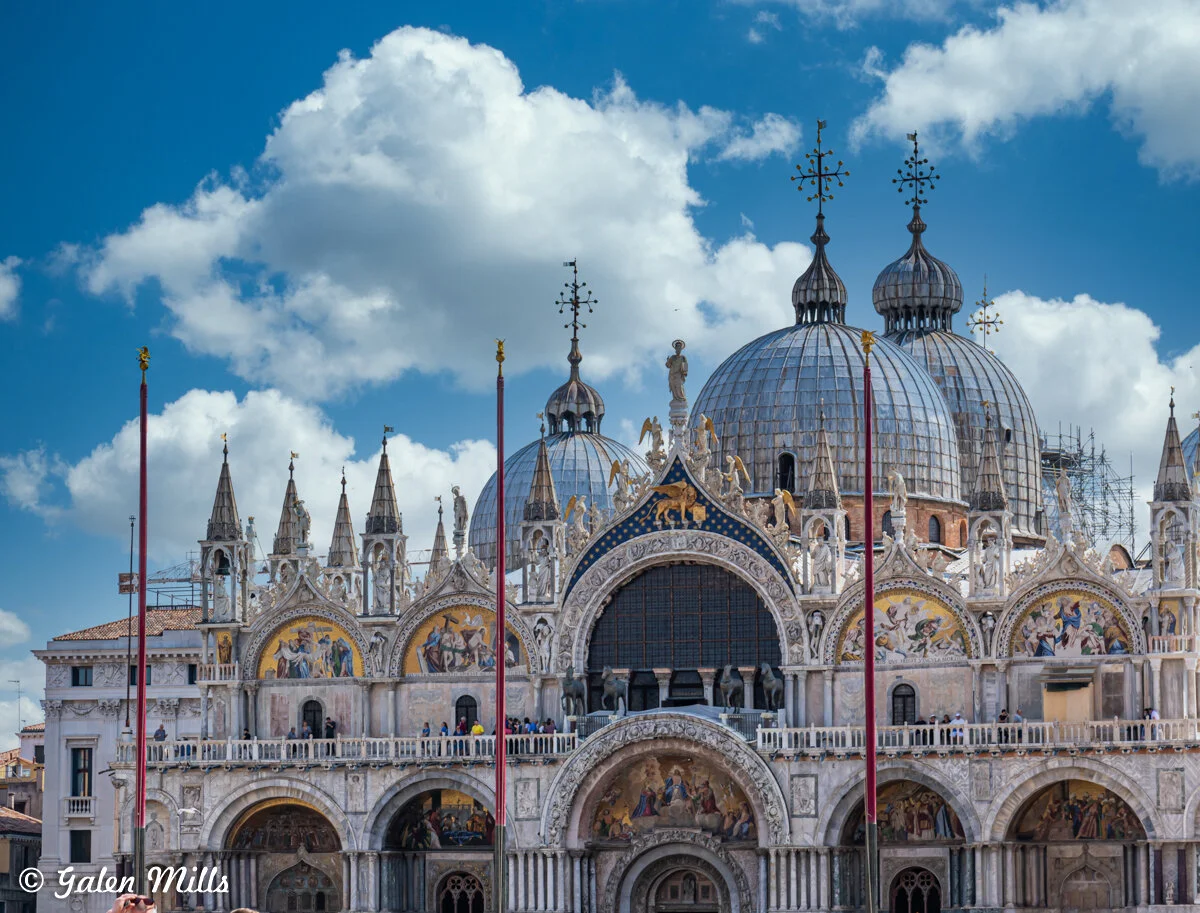 St. Mark's Basilica in Venice, with ornate facade, domes, and spires, set against a blue sky with clouds.
